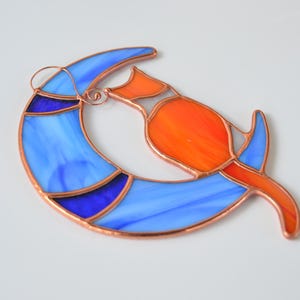 May include: A stained glass suncatcher depicting a stylised orange cat curled up on a blue crescent moon. The cat and moon are outlined in copper.