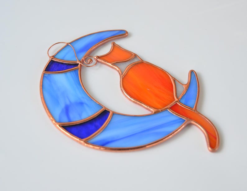May include: A stained glass suncatcher depicting a stylised orange cat curled up on a blue crescent moon. The cat and moon are outlined in copper.