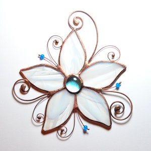 Peut inclure: Une fleur en verre blanc avec un bijou bleu au centre. La fleur est entour&eacute;e de fil de cuivre avec des volutes.