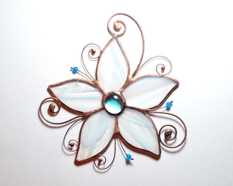 Peut inclure: Une fleur en verre blanc avec un bijou bleu au centre. La fleur est entour&eacute;e de fil de cuivre avec des volutes.