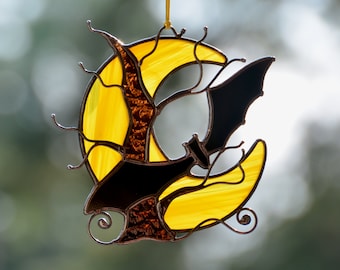 May include: A stained glass suncatcher depicting a yellow crescent moon and a black bat. The moon's centre is textured and amber-coloured, with a dark metal outline. A small loop is attached for hanging, suitable for Halloween.