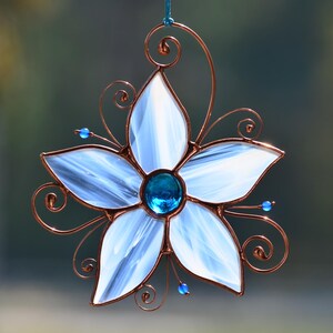 Peut inclure: Une fleur en verre blanc avec un bijou bleu au centre, entour&eacute;e de volutes de fil de cuivre. La fleur est suspendue &agrave; une boucle de fil de cuivre.