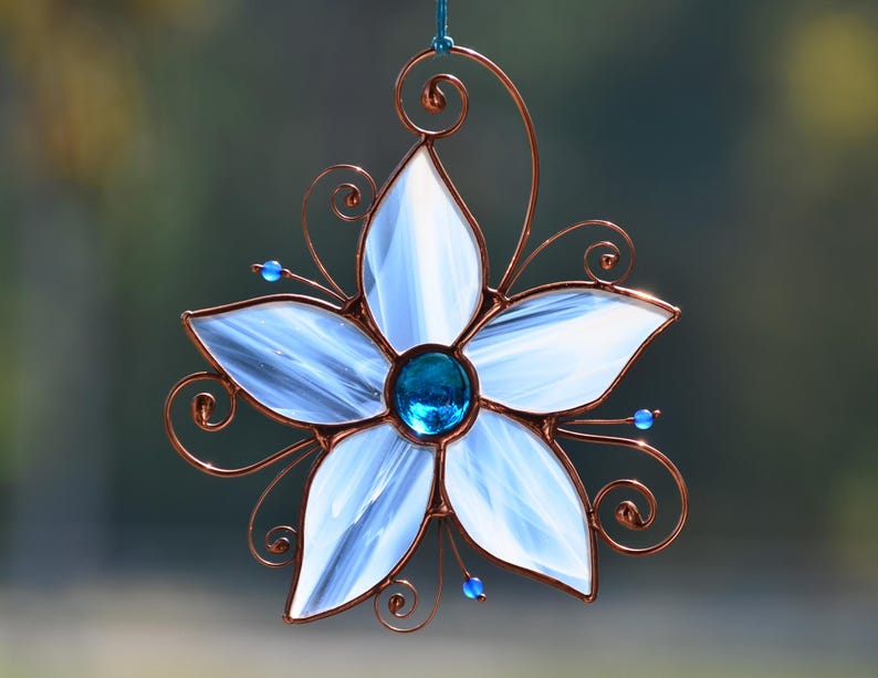 Peut inclure: Une fleur en verre blanc avec un bijou bleu au centre, entour&eacute;e de volutes de fil de cuivre. La fleur est suspendue &agrave; une boucle de fil de cuivre.