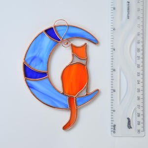 May include: A stained glass suncatcher depicting a cat sitting on a crescent moon. The moon is blue with white and blue swirls. The cat is orange with white accents.