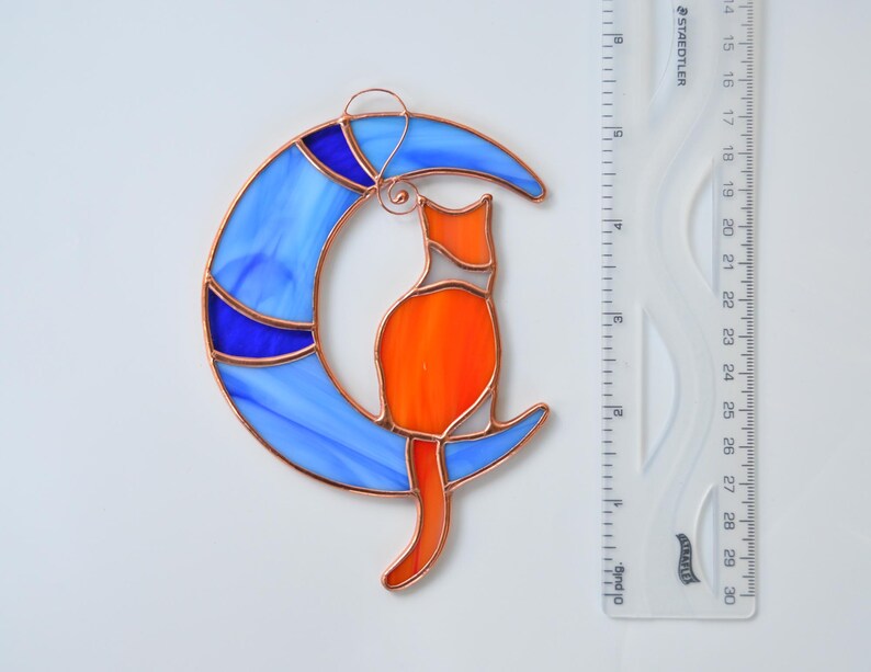 May include: A stained glass suncatcher depicting a cat sitting on a crescent moon. The moon is blue with white and blue swirls. The cat is orange with white accents.