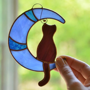Puede incluir: Un atrapasol de vidrio coloreado con una luna creciente azul con un dise&ntilde;o azul y turquesa y un gato marr&oacute;n sentado en la luna.