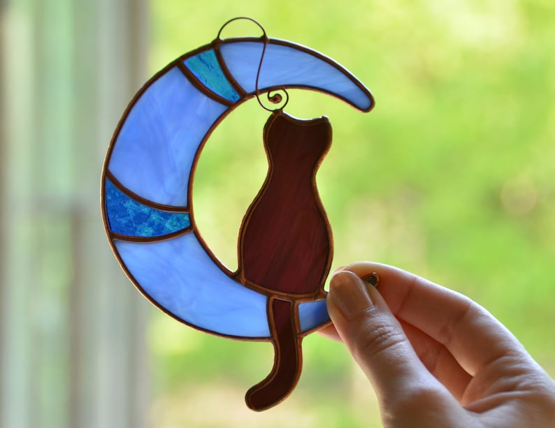Puede incluir: Un atrapasol de vidrio coloreado con una luna creciente azul con un dise&ntilde;o azul y turquesa y un gato marr&oacute;n sentado en la luna.