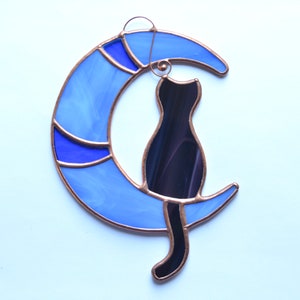 Puede incluir: Una ventana de vitral colgante con una silueta de gato negro sentada en una luna creciente azul.