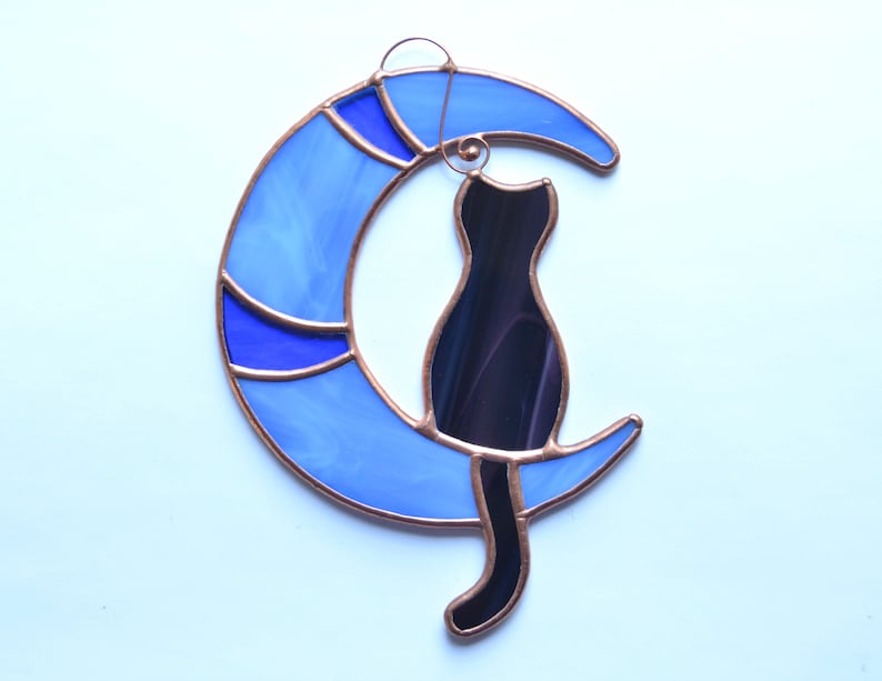 Puede incluir: Una ventana de vitral colgante con una silueta de gato negro sentada en una luna creciente azul.