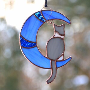 May include: A stained glass cat sitting on a blue crescent moon. The cat is grey with white markings. The moon has a blue and white design.