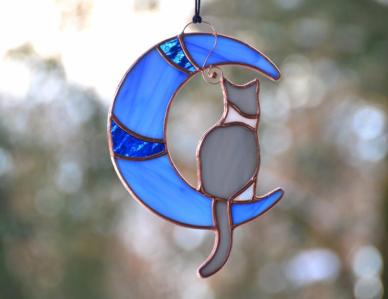 May include: A stained glass cat sitting on a blue crescent moon. The cat is grey with white markings. The moon has a blue and white design.