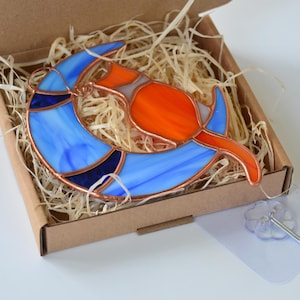 May include: A stained glass suncatcher depicting a cat sitting on a crescent moon. The moon is blue with white swirls and the cat is orange with white accents.