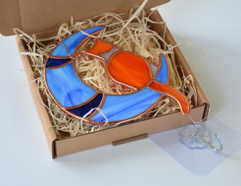 May include: A stained glass suncatcher depicting a cat sitting on a crescent moon. The moon is blue with white swirls and the cat is orange with white accents.