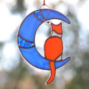 May include: A stained glass suncatcher depicting a red cat sitting on a blue crescent moon. The cat has white markings on its face and chest. The moon has a blue and white swirl pattern.
