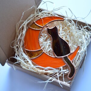 Puede incluir: Un gato de vidrio coloreado sentado en una luna creciente naranja. El gato es negro y la luna tiene un contorno de cobre.