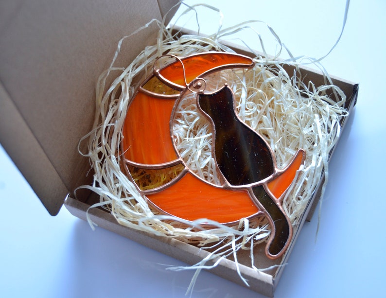 Puede incluir: Un gato de vidrio coloreado sentado en una luna creciente naranja. El gato es negro y la luna tiene un contorno de cobre.