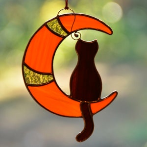 Puede incluir: Un atrapasol de vidrio coloreado con un gato negro sentado en una luna creciente. La luna es naranja con detalles amarillos.
