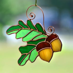 May include: A stained glass ornament depicting a green oak leaf with two acorns. The acorns are brown and yellow with copper wire accents.