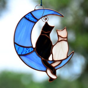 May include: A stained glass suncatcher depicting two cats on a blue crescent moon. One cat is black, the other white. The moon has a blue and light blue design with copper borders and a hanging loop.