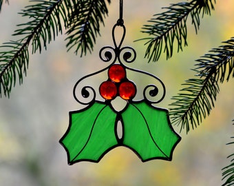 May include: A stained glass holly ornament with red berries and green leaves. The ornament is hanging from a branch.