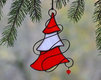 May include: A stained glass Christmas tree ornament with red and white sections, outlined in black wire. A small red bead hangs from the bottom. The ornament is suspended from a black cord against a blurred background.