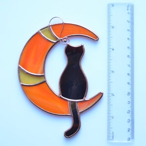 Puede incluir: Un gato de vidrio coloreado sentado en una luna creciente. La luna es naranja y amarilla con un contorno de cobre. El gato es negro con un contorno de cobre.