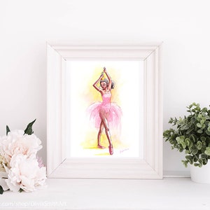 May include: Watercolor painting of a ballerina in a pink tutu and pointe shoes, arms raised. The artwork is framed in a white wooden frame. The background is yellow and the ballerina is in a dancing pose. The artwork is displayed on a white surface.