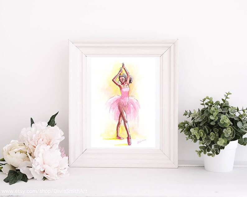 May include: Watercolor painting of a ballerina in a pink tutu and pointe shoes, arms raised. The artwork is framed in a white wooden frame. The background is yellow and the ballerina is in a dancing pose. The artwork is displayed on a white surface.