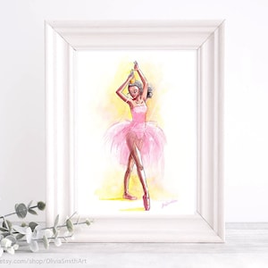 May include: Watercolor painting of a ballerina in a pink tutu and pointe shoes, framed in a white wooden frame. The ballerina is depicted with her arms raised, wearing a crown. The background is a soft yellow and orange wash.