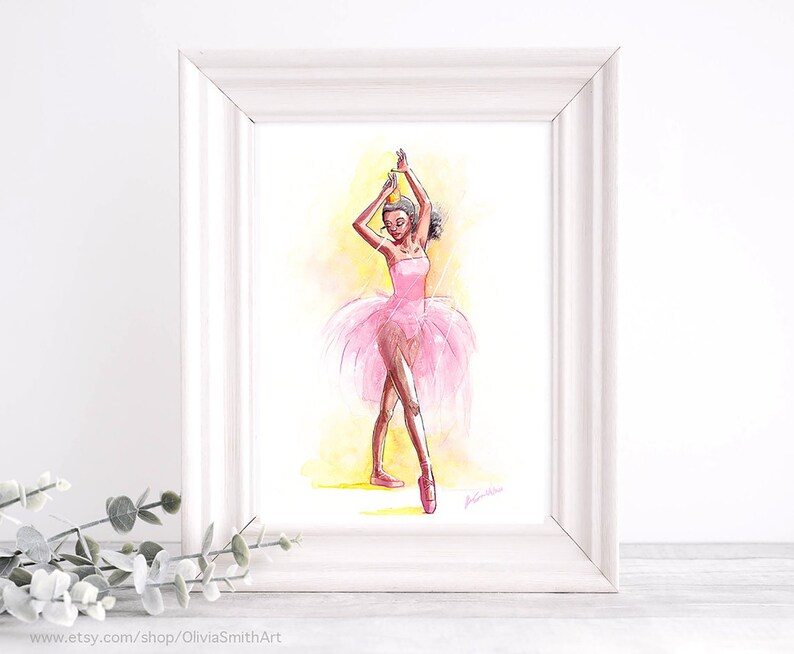 May include: Watercolor painting of a ballerina in a pink tutu and pointe shoes, framed in a white wooden frame. The ballerina is depicted with her arms raised, wearing a crown. The background is a soft yellow and orange wash.