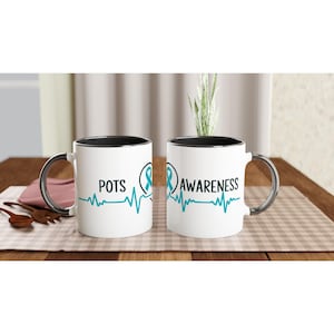 May include: Two white ceramic mugs with black interiors and silver handles. One mug reads "POTS" and the other reads "AWARENESS" in black text, with a teal ribbon and heartbeat line design. The mugs are on a wooden table.