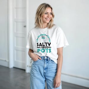 Pode incluir: Camiseta branca com o texto "I'M ALLOWED TO BE SALTY I HAVE POTS" em preto e verde-azulado. O gráfico inclui uma ilustração de um saleiro. A pessoa está usando jeans azul claro.