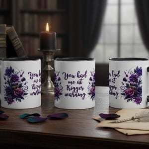 Può includere: Tre tazze da caffè bianche con manici e bordi neri. Ogni tazza presenta un motivo floreale con rose viola e la scritta "You had me at trigger warning". Una candela e dei libri sono sullo sfondo.