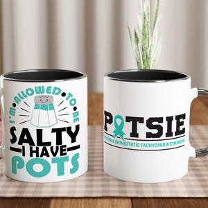 Peut inclure: Deux mugs en céramique blanche avec des bords noirs et des poignées argentées. L'un des mugs indique "I'M ALLOWED TO BE SALTY I HAVE POTS" avec un graphique de salière. L'autre mug dit "POTSIE" avec un ruban turquoise et du texte sur le syndrome de tachycardie orthostatique.