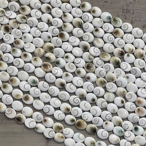 May include: A strand of white oval-shaped shell beads with brown and green swirls. The beads are arranged in rows and are on a light brown surface.