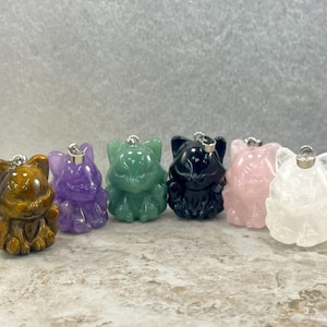 May include: Six small, carved cat figurines made of different types of gemstones. The cats are all in a sitting position with their paws tucked under them. The gemstones are tiger's eye, amethyst, green aventurine, black obsidian, rose quartz, and clear quartz.