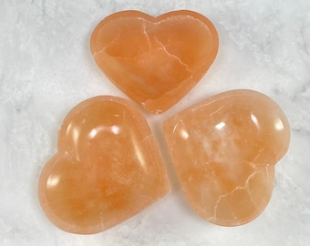 May include: Three heart-shaped orange calcite crystal bowls. The bowls are smooth and polished, with a natural, slightly translucent appearance.