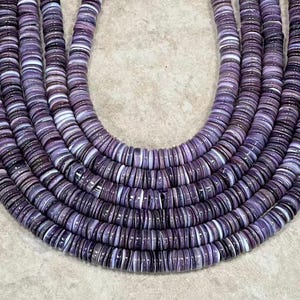 May include: Close-up of several strands of purple and white disc-shaped beads. The beads are arranged in a curved pattern, showcasing their varying shades of purple and white. The beads appear to be made of a natural material, possibly shell or stone.