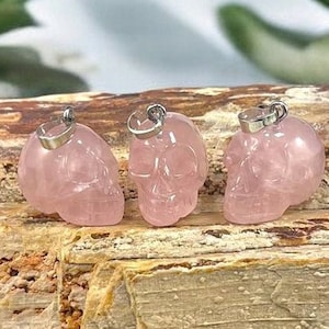 Pode incluir: Três pingentes de caveira de cristal rosa com argolas de prata. As caveiras são esculpidas em quartzo rosa e estão sobre um pedaço de madeira petrificada.