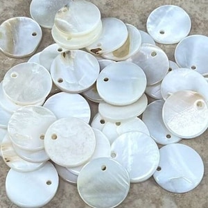Puede incluir: Un montón de discos redondos de nácar blanco con un agujero en el centro. Los discos tienen un diámetro de aproximadamente 2,5 cm.