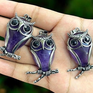 May include: Three silver owl pendants with amethyst stone insets and black onyx eyes. The owls are perched on a hand with a blurred background of green foliage.