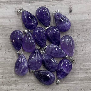 May include: A group of 13 amethyst teardrop pendants with silver bail settings. The pendants are arranged in a pile on a light gray textured surface.