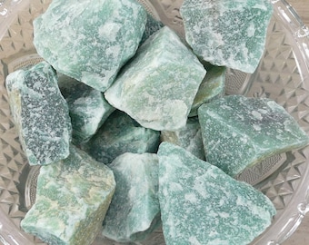 May include: A collection of rough, green aventurine stones in a clear glass bowl. The stones vary in size and shape, with a textured, crystalline appearance. The color ranges from pale to deep green, with some areas appearing almost white.