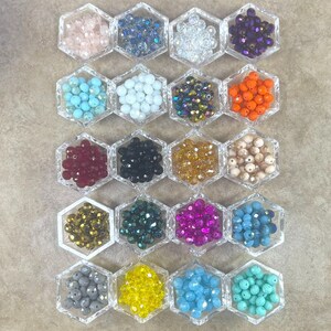 Könnte beinhalten: Eine Sammlung facettierter Perlen in verschiedenen Farben, darunter Rosa, Blau, Weiß, Lila, Türkis, Orange, Rot, Schwarz, Gold und Beige. Die Perlen werden in transparenten, sechseckigen Behältern präsentiert, ideal für Schmuckherstellung oder Bastelprojekte.