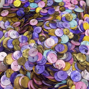 May include: A collection of colorful, round buttons in various shades of pink, purple, yellow, and gold. The buttons have two or four holes for sewing and are made of a shiny, iridescent material. The buttons are scattered in a pile.