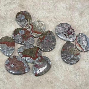 May include: A collection of polished, teardrop-shaped stones. The stones feature a mix of colors, including gray, red, and brown, with unique patterns and textures. The stones are arranged on a light-colored surface.