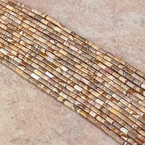 Può includere: Una collezione di perline di pietra naturale nei toni del beige, marrone e crema. Le perline sono cilindriche, con una forma leggermente irregolare, e sono infilate insieme. Le perline hanno un aspetto screziato, con variazioni di colore e motivo.
