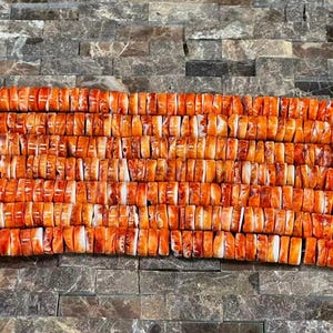 May include: Close-up of a strand of orange and white spiny oyster shell beads. The beads are rectangular and have a natural, organic appearance. The beads are strung together, creating a textured and colorful pattern. The background is a dark, textured surface.