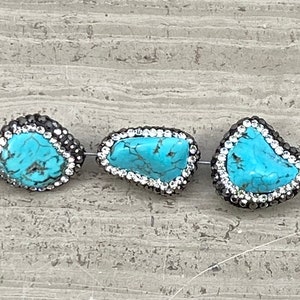 May include: Three turquoise gemstone beads with silver accents and rhinestones. The beads are irregularly shaped and have a natural, organic look.