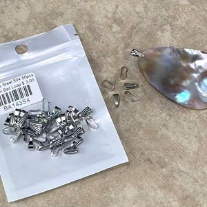 Puede incluir: Una bolsa de plástico transparente que contiene 50 bucles de fianza a presión de acero inoxidable. Junto a la bolsa hay un colgante nacarado con una fianza plateada y varios bucles de fianza a presión sueltos. La bolsa tiene una etiqueta que dice "Stainless Steel 304 50pcs Snap On Bail Loop".